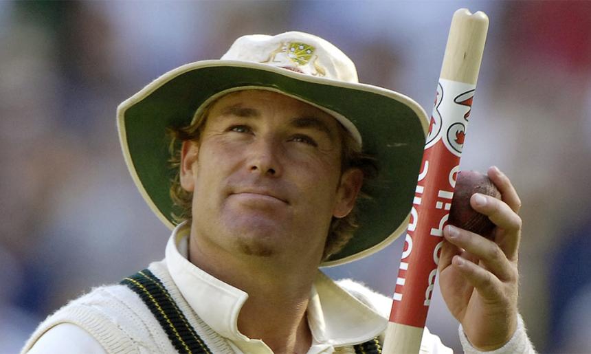 A man in cricket whites and a hat holding a cricket stump and looking upwards.