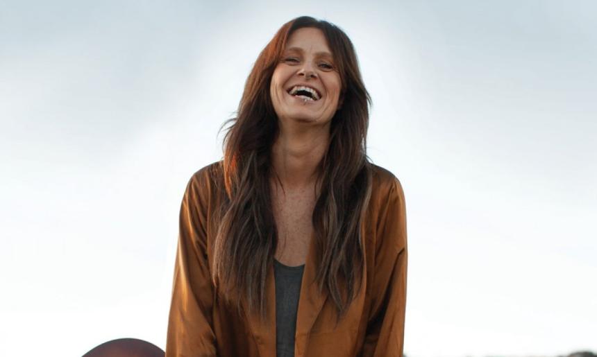 A woman with long brown hair, wearing a brown shirt and holding a guitar and smiling.