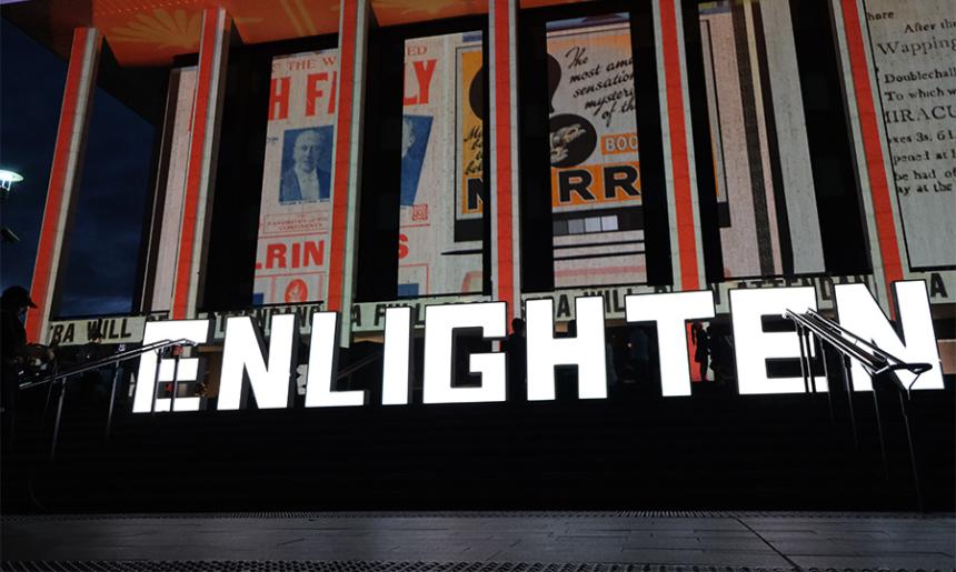 The front of the National Library of Australia, lit up with images and lights. There are big light up letters in front of the building that spell out 'Enlighten'.