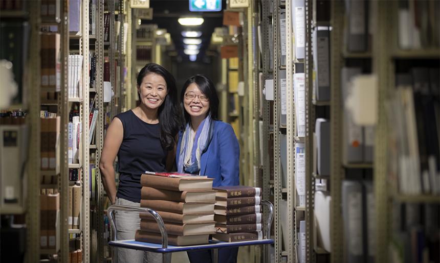 Two women standing behind a trolley with books on it in the Library stacks.