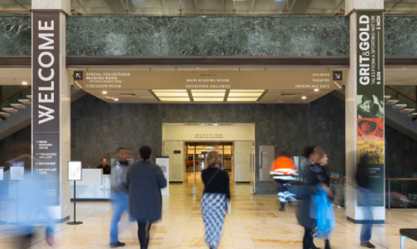 Large two-story room with cream-coloured marble floors, columns and stairs; black leather couches; lots of light from the ceiling and a balcony from the second floor looking over the room. On the columns are signs with directions and the word 'Welcome'. People are moving around the room