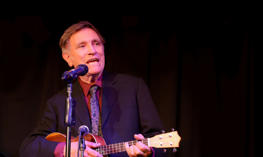 Man in a suit and tie playing guitar and singing behind two microphones