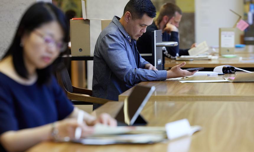 Three people sitting at wooden tables, looking at National Library collection material.