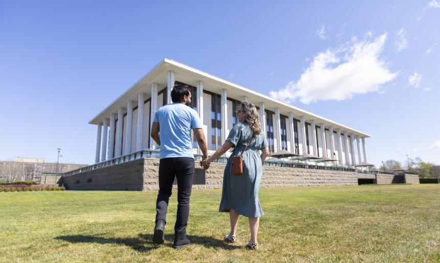 Man and woman in casual wear holding hands and laughing as they walk towards the National Library building