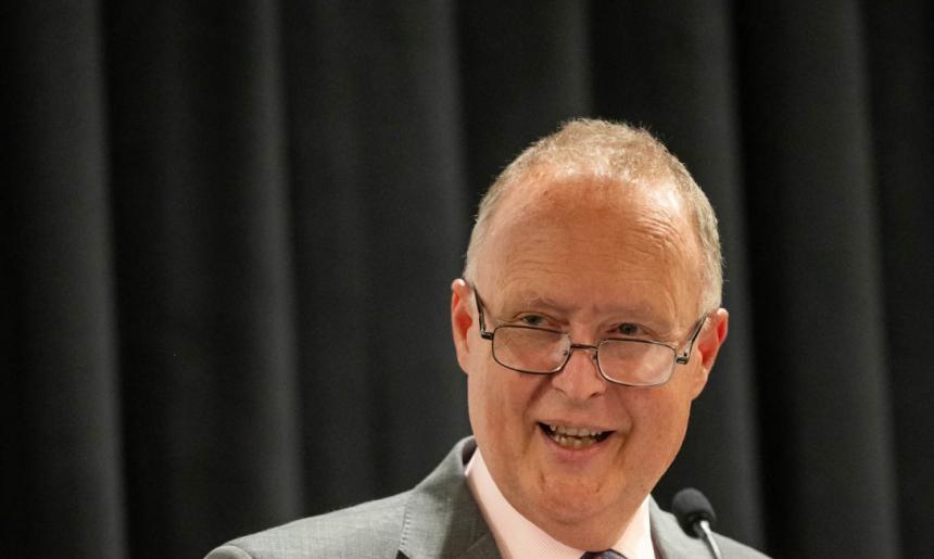 Rupert Myer AO, wearing a grey suit with a pink shirt and patterned tie, speaks at a podium with a microphone. He is smiling warmly and wearing glasses, addressing an audience against a backdrop of dark curtains.