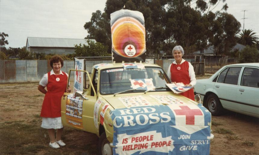Two ladies in nurses uniforms standing either side of an old car that is covered in Red Cross logos and images.
