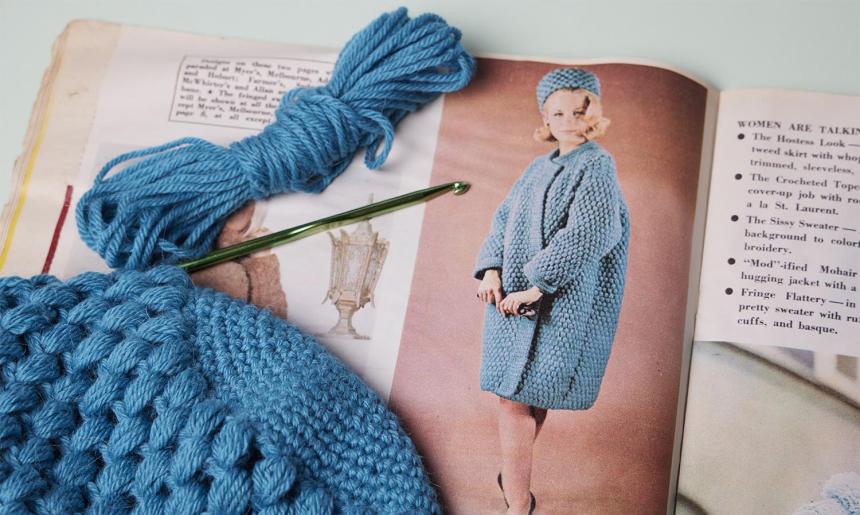 An old magazine showing a vintage crochet pattern. Laying on top of the magazine is a blue crochet item in progress.