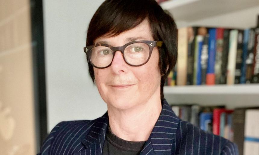 A portrait image of a person with short hair, wearing glasses and a dark blue striped blazer. They are standing in front of a bookshelf.