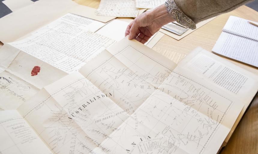 Hand holding up the top corner of a map of Australia, on a table filled with other map and manuscript material