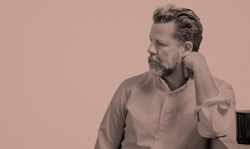 An image of a man leaning on one arm and looking to the left. The image has a pink colour wash over it.