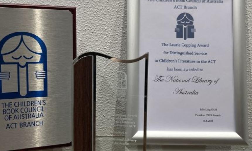 A silver and wooden plaque, glass trophy and framed certificate for the 2024 CBCA Laurie Copping Award given to the National Library