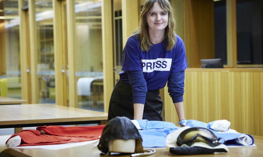 Dr Jo Langdon standing and leaning on a table in the Special Collections Reading Room with a Royal Perth Hospital Nurses uniform worn including a red cape and two pilot's helmets and goggles on it