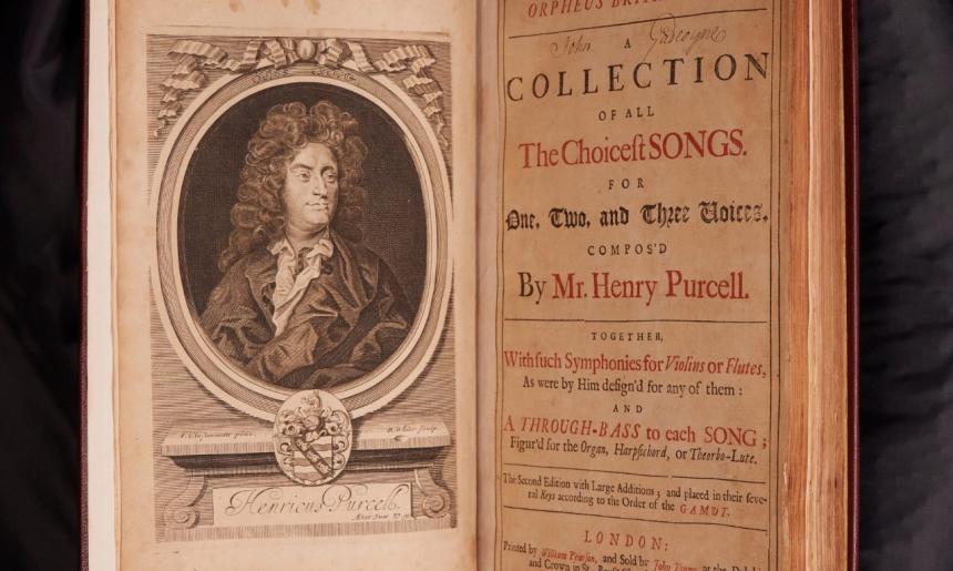 Old book sitting open on a black cushion. On the left page is a portrait of a man with large rolled hair, on the right is the title page with text reading 'Orpheus Britannicus; A collection of all the choicest songs for one, two and three voices, composed by Mr. Henry Purcell'