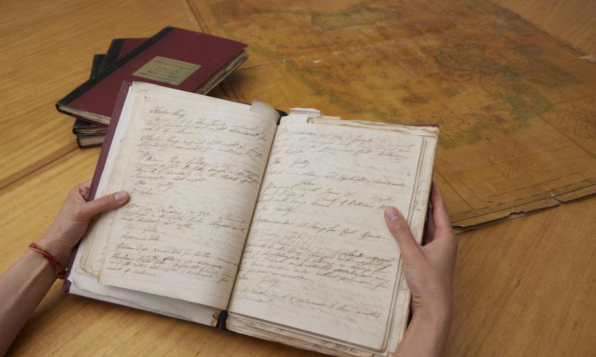 Hand holding an old notebook with handwritten text. In the background is a stack of books and an old map.