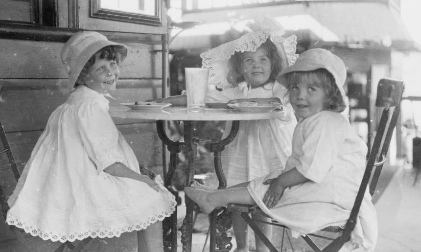 Three young girls in white summer dresses and hats sitting at a cafe table