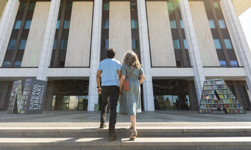 Man and woman in casual wear holding hands and laughing as they walk up the stairs towards the National Library entrance