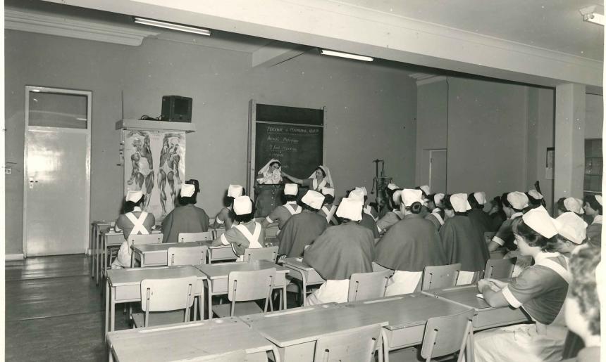 Black and white photo of a classroom full of women in nursing uniforms listening to the teachers at the front of the room who are standing in front of a blackboard and near an anatomical chart