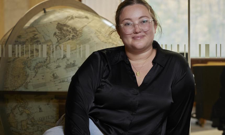 Woman, Jacinta Mackay, sitting in front of a window looking into the Main Reading Room with a large globe on the other side