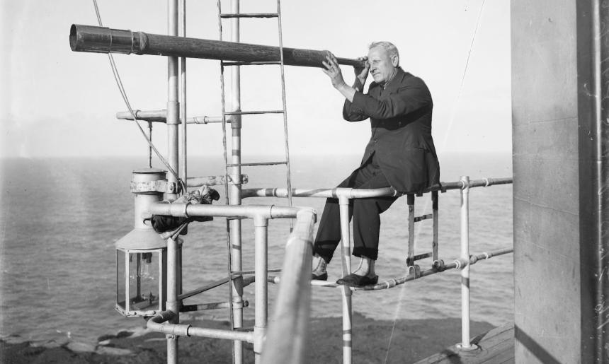 Black and white photo of a man sitting on a balcony railing near the ocean, looking into a very long telescope