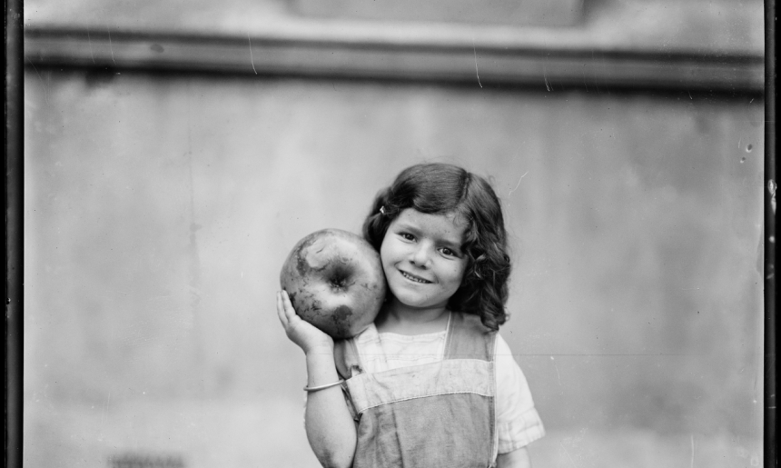 Black and white image of a young girl smilng and holding a large apple up on her shoulder