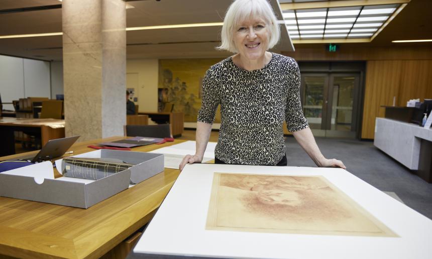 Woman, Dr Christina Thompson, standing in the Special Collections Reading Room, leaning on a table with a large portrait on it and smiling