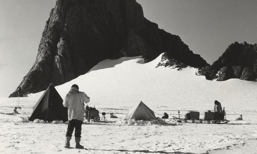 A man standing on a snowy plain, in front of tents, sledges with scientific equipment, scientists and sled dogs. A large rocky outcrop looms in the background. 