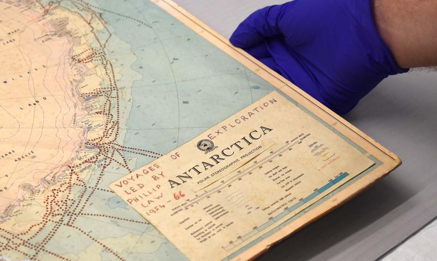 A hand wearing a blue glove holds the bottom right corner of a map of Antarctica including the handwritten words 'Voyages of Exploration Led By Phillip Law 1954-66'