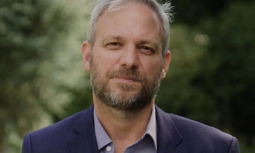 A man with grey hair and brown and grey beard and moustache standing in front of blurred out trees