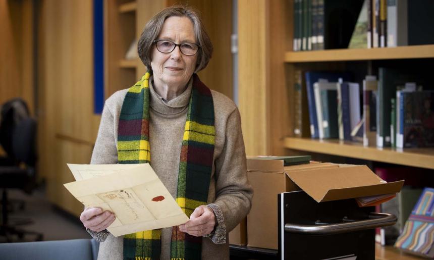 A woman with short hair and glasses smiles and looks at the camera while she holds a folded document in her hands. In the background are bookshelves with books and an open box on a trolley.