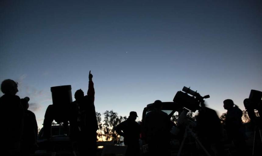 A group of people at dusk standing beside small telescopes and pointing at the sky.