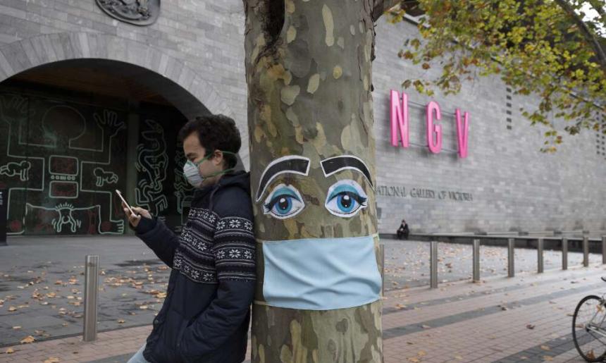 A man wearing a face mask leaning against a tree that is decorated with a face mask and eyes outside the NGV in Melbourne