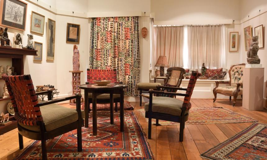 A cosy living room. A small table surrounded by four chairs takes up much of the image. There are a few other seats in the image, and artwork and sculpture adorning the walls and shelves lining the walls.