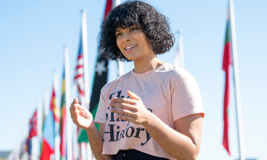 A women wearing a pink t-shirt with the words 'She Shapes History' delivers a guided tour in front of several flagpoles