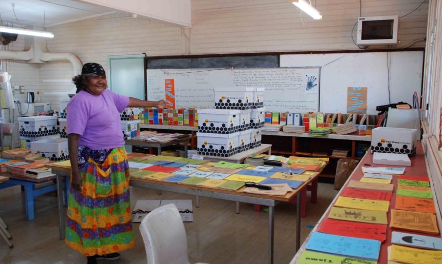 A woman stands in a classroom gesturing at piles of readers laid out on the tables in the classroom.