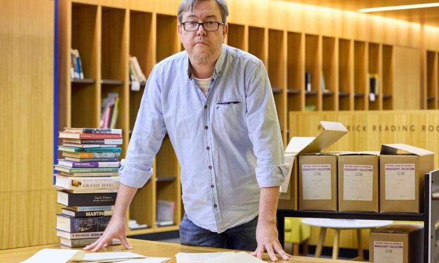 A man in a blue shirt leaning over a desk with papers and books