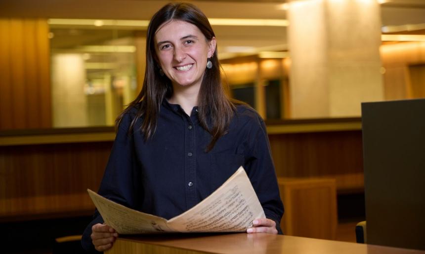 A smiling woman with long dark hair is seated at a desk, looking directly at the camera while holding and reading a paper.
