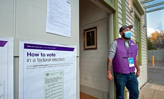 Man wearing face mask and purple vest standing new a doorway. On the wall around the door are instructions on how to vote in a federal election.