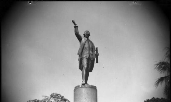 A black-and-white photograph of a statue of Captain Cook on a tall pedestal. The statue shows Cook standing with one arm raised and holding a rolled document in the other hand. Trees and part of a lamppost are visible in the foreground.