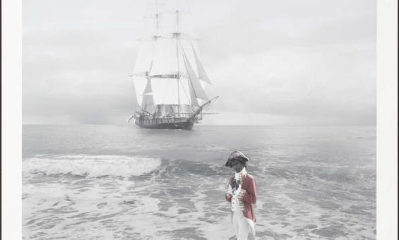 An Aboriginal person dressed in a red coat with white breeches and black boots stands on a beach, just at the edge of the water. They wear a tricorn hat with a red naval officer jacket and holds a scroll. In the background, a large, multi-masted sailing ship with white sails is anchored on calm waters under an overcast sky.