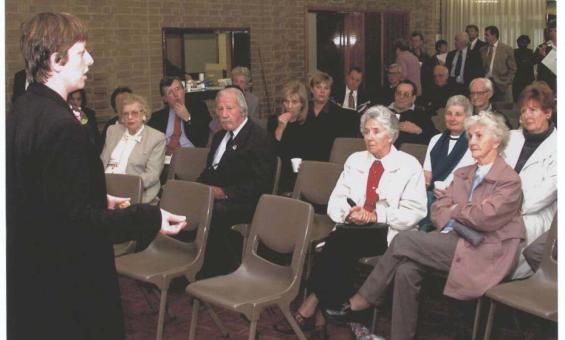 A photo of a woman standing and speaking to a room of seated and standing people.