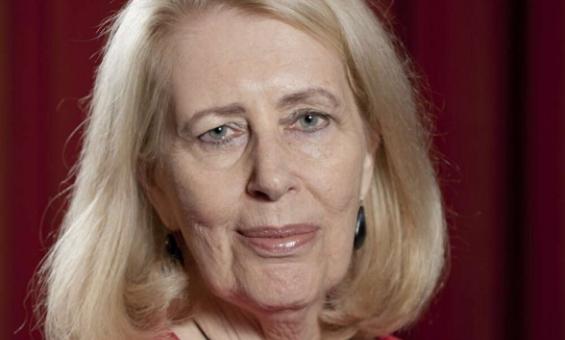 A colour photograph of Anne Summers. Anne has shoulder length blonde hair and wears a red outfit and a large round necklace, standing against a red curtain background.