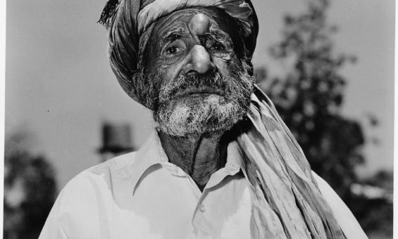 A black-and-white portrait of an older man with a weathered face and a beard, wearing a turban and a white shirt. He stands outdoors, with blurred trees in the background.