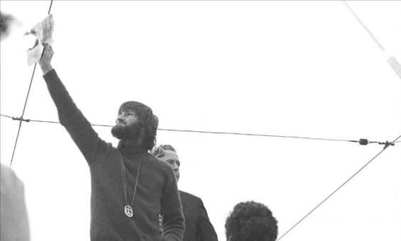 A black and white photo of a bearded man with collar length hair and a large metal peace medallion hanging down his front, holds a burning piece of paper up high, while several people watch and take photos.