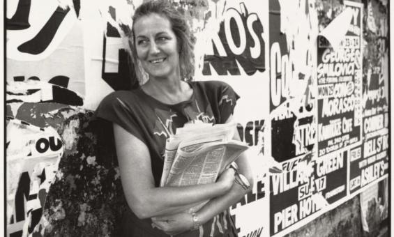 A black-and-white photograph of a woman standing against a wall covered in posters. She is smiling and holding a folded newspaper in her arms, casually dressed with loose hair. The posters in the background are partially torn and feature bold text and graphics.