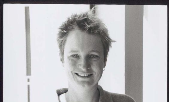 A black and white portrait photo of a young, freckle faced, smiling woman with very short, wild, fair hair, holding a book open.