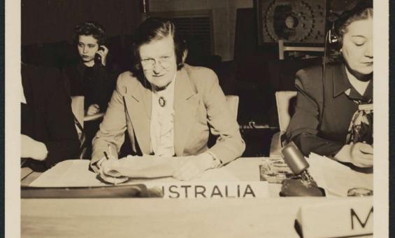 A black-and-white photograph of Jessie Street seated at a desk representing Australia at a United Nations conference. She is wearing a white shirt and suit jacket and is writing, with other women visible beside her.
