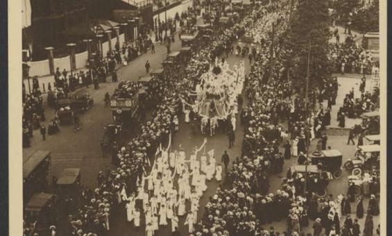 An aerial view of a large street procession with participants dressed in white, marching in rows and carrying banners. The crowded scene is framed by towering buildings on one side and tree-lined streets on the other. Spectators line the sidewalks, watching the organised march as cars and horse-drawn carriages are scattered among the crowd. 