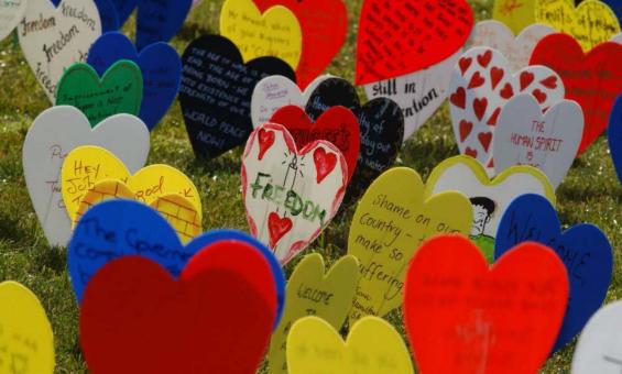 A photo of lots of different coloured plastic love hearts on a wire, stuck in a grassy area. The hearts are decorated with writing about freedom, and drawings.