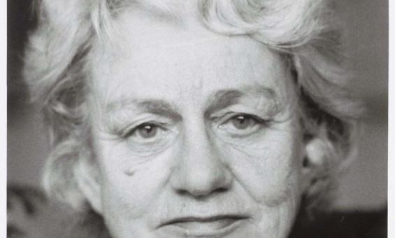 A black and white portrait photo of an elderly woman with short white, wavy hair wearing a black roll neck jumper and a small floral brooch just below the collar, with her interlaced fingers resting in the foreground.