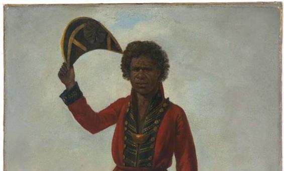 A portrait of a man. He has dark skin and hair. He is an Australian Indigenous man and is wearing a European style officers coat and trousers. His trousers are tattered at the cuffs. He is holding a tricorn hat in the air. In the background sailing ships can be see on the harbour. A large fort can be seen on a spit of land.